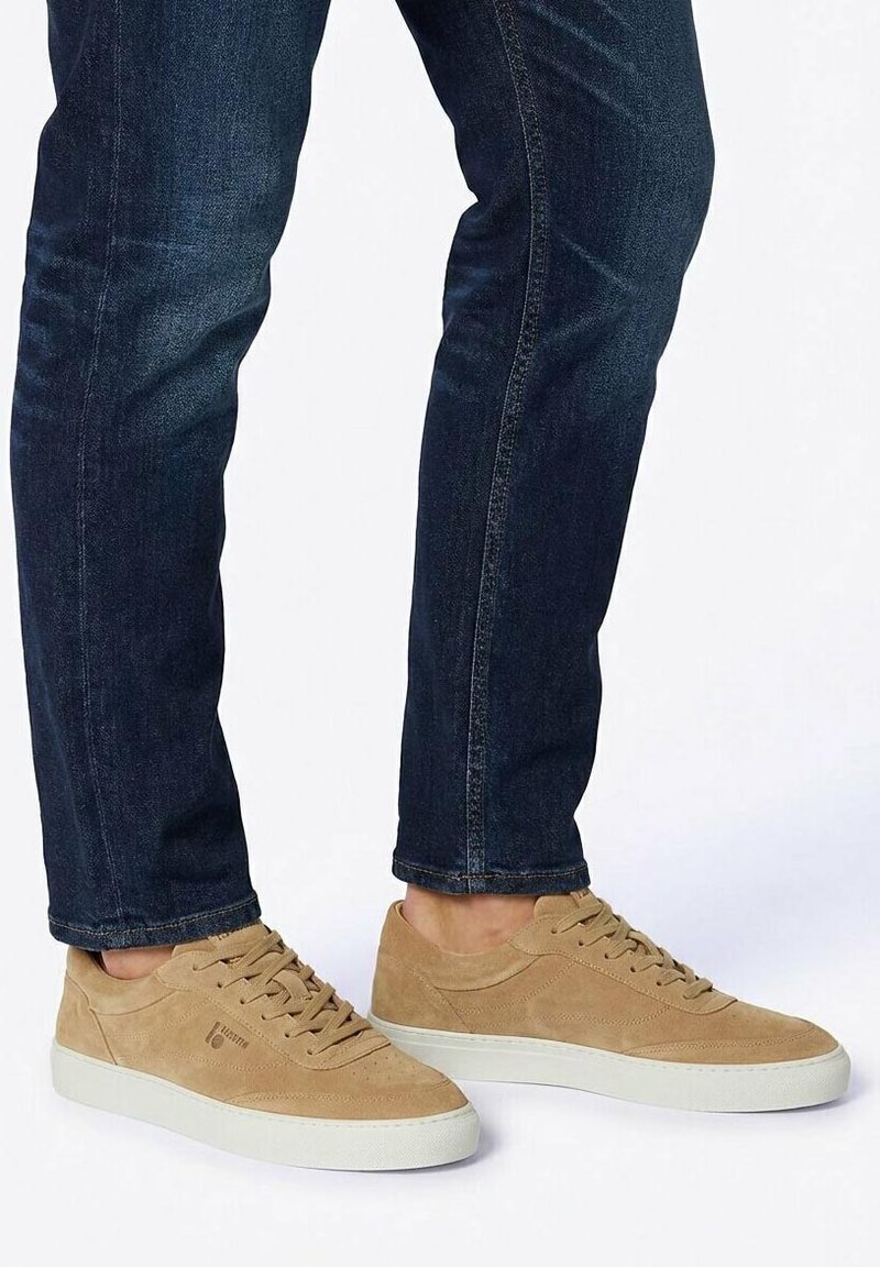 Persoon die donkerblauwe spijkerbroek en beige suède sneakers met witte zolen draagt, staand op een wit oppervlak.