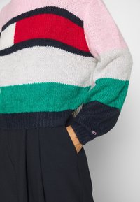 Farverig strikket sweater med horisontale striber i pink, rød, hvid, grøn og marineblå, samt struktureret stof og logo-detalje på ærmet.