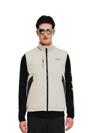 Leichter Creme-Faux-Down-Westen mit hohem Kragen, schwarzen Akzenten, Reißverschluss und Logo auf der Brust. Das Modell trägt ein schwarzes Langarmshirt darunter.