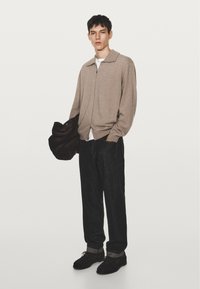 Ung man i beige zip-up tröja, svarta jeans, svarta skor, som håller en mörkbrun väska, stående mot en enkel vit bakgrund.