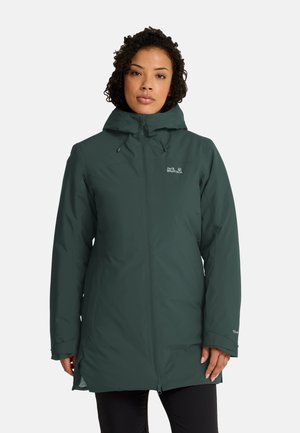 Femme portant une longue doudoune d'hiver vert foncé à capuche avec une fermeture éclair intégrale à l'avant et un logo sur la poitrine, debout sur un fond uni.