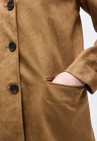 Κλείσιμο φωτογραφίας ενός χεριού που μπαίνει μερικώς στην τσέπη ενός καφέ σουέτ παλτού με τρία μαύρα κουμπιά.