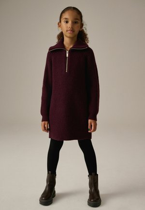 Robe pull tricotée en bourgogne profond avec un col montant et une fermeture éclair sur le devant, manches longues et texture côtelée. Portée sur des collants noirs et des bottines.