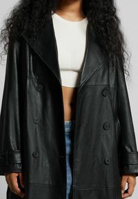 Abrigo de cuero negro de doble botonadura con grandes solapas, detalles de botones y una textura suave. Presenta un corte relajado y mangas anchas.