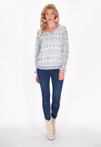 Blusa bianca leggera con motivo ikat blu, scollo a V e maniche lunghe. Abbinata a jeans blu scuro e stivali da cowboy beige.