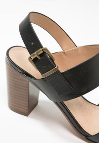 Sandale à talon haut en cuir noir avec une large sangle, une boucle réglable et un talon en bois texturé brun. Doublure intérieure lisse.