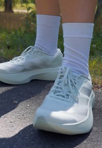 Scarpe da atleta verde chiaro con pannelli in rete, superfici texture e lacci piatti, abbinate a calzini bianchi alla caviglia, su un sentiero asfaltato.