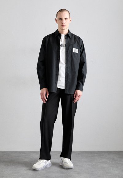 Black button-up shirt, lightweight fabric, featuring a chest pocket with a white barcode label. Paired with black trousers and white sneakers.