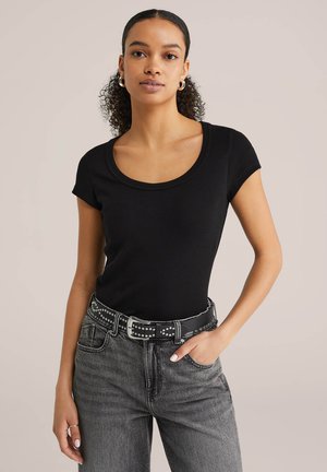 Göndör hajú nő fekete, testhezálló rövid ujjú felsőben, szegecses fekete övvel és szürke farmernadrágban, egyik kezét a zsebében tartva áll.