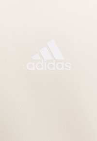 Biała tkanina sportowa z teksturowaną powierzchnią. Posiada wyraźne białe logo Adidas z trzema ukośnymi paskami nad nazwą marki.