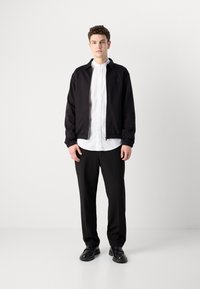 Schwarze Bomberjacke über einem weißen Hemd, schwarzen Hosen und schwarzen Schuhen. Glattes Material, gerippte Bündchen und dezentes Logodetail auf der Jacke.