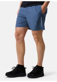 Blå sportshorts i lättviktig tyg med en slät yta; har en sidoficka med reflexdetalj. Bärs med svarta sneakers.