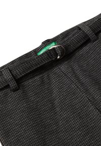 Detailní záběr na tmavě šedé látkové kalhoty s texturou, s nastavitelným látkovým opaskem a malou zelenou etiketou se nápisem "UNITED COLORS."