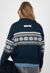 Personne portant un pull bleu marine avec des motifs de flocons de neige blancs dans le dos et sur les manches, tenant des vêtements foncés dans une main.