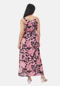 Donna con lunghi capelli scuri che indossa un abito maxi senza maniche floreale nero e rosa e sandali con tacco trasparente, in piedi con la schiena rivolta.