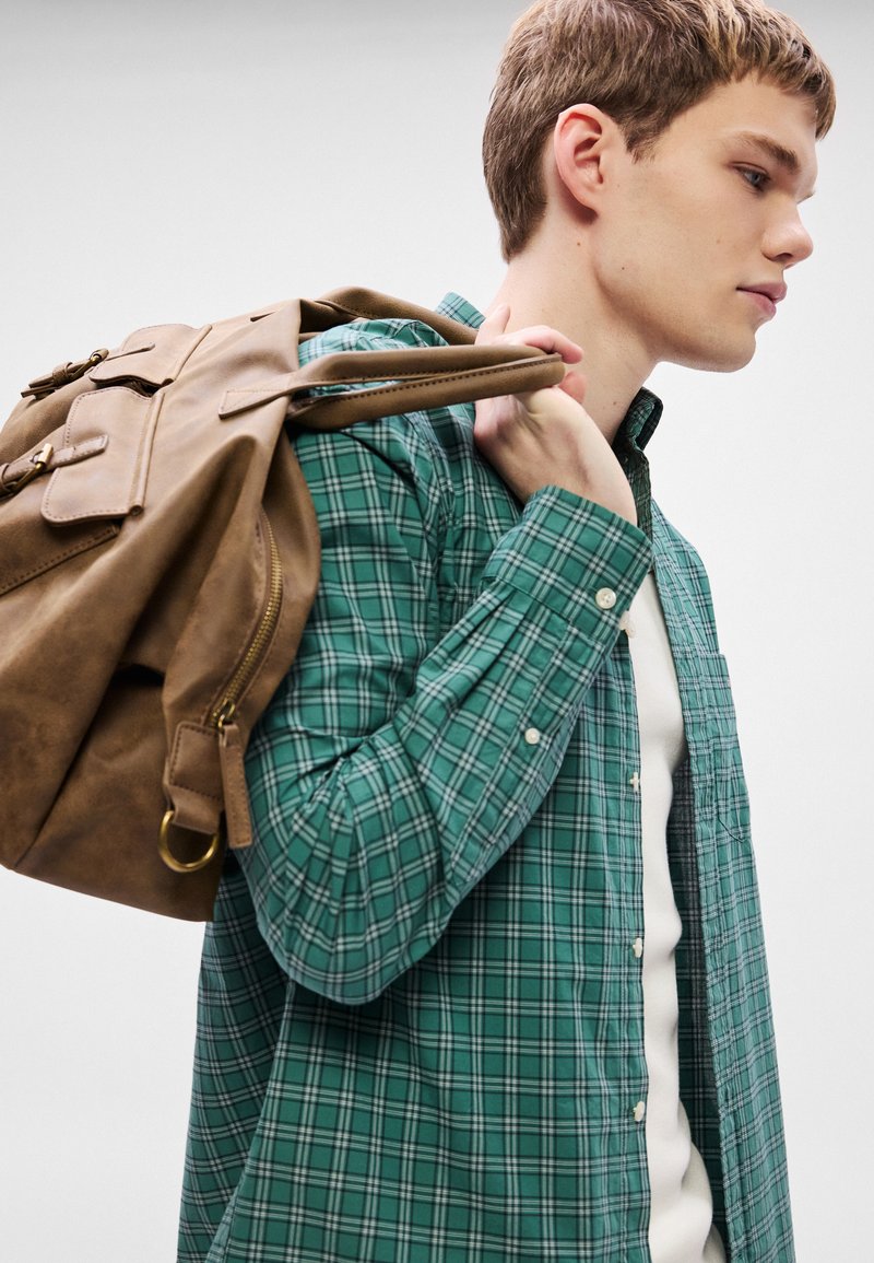 Green plaid shirt over a white top, worn by a person carrying a brown, soft-textured backpack with zippers and flat pockets.