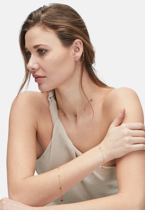 Goldfarbene, geschichtete Schmuckstücke mit einer zarten Halskette mit Charms und einem verstellbaren Armband. Das Model trägt ein leichtes, strukturiertes Oberteil.
