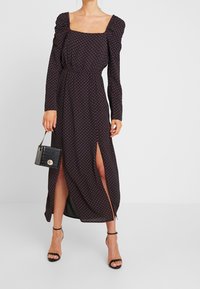 Lång svart klänning med långa ärmar, fyrkantig halsringning, röda prickar, slitsar på sidorna och åtsittande midja; kombinerad med en liten svart handväska.