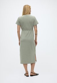 Γυναίκα με ξανθά μαλλιά που φοράει midi φόρεμα με πράσινες και λευκές ρίγες, κοντά μανίκια, πλαϊνή σκίσιμο και μαύρα επίπεδα παπούτσια, με την πλάτη γυρισμένη.