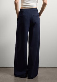 Personne portant un pantalon navy taille haute et large ainsi qu'un haut court blanc, se tenant contre un mur clair et uni avec les mains dans les poches.