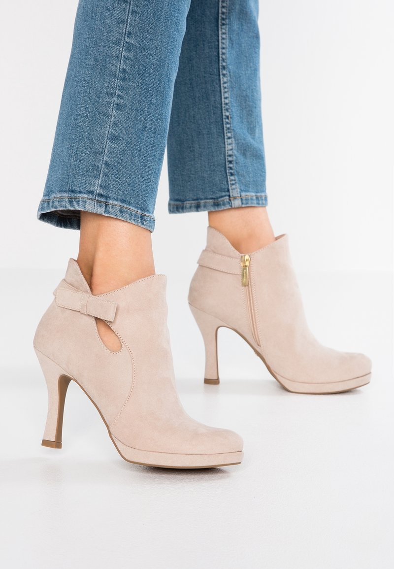 Bottines beiges en daim à talons hauts avec fermeture éclair latérale et détail nœud, portées avec un jean court bleu sur fond blanc.