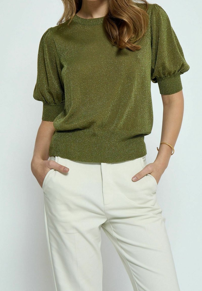 Pull en maille vert olive scintillant avec manches bouffantes et ourlet côtelé, associé à un pantalon beige clair. Texture lisse et coupe ajustée.