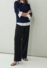 Pull marin en tricot avec ourlet et épaules côtelés, superposé sur une chemise blanche à col, associé à un pantalon large noir et des sandales noires.