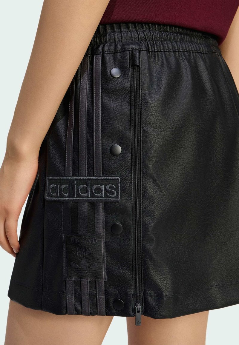 Woman wearing a black faux leather Adidas skirt with side zipper, snap buttons, and branded stripes on the left hip.