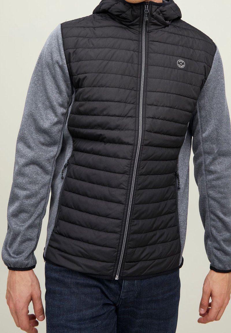 Black and gray zip-up jacket with quilted body, smooth sleeves, a hood, and side pockets. Features a logo on the chest.