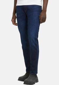 Jeans in denim blu scuro con design a gamba dritta, caratterizzati da dettagli di cucitura arancione e una texture liscia, abbinati a stivali alla caviglia grigi.
