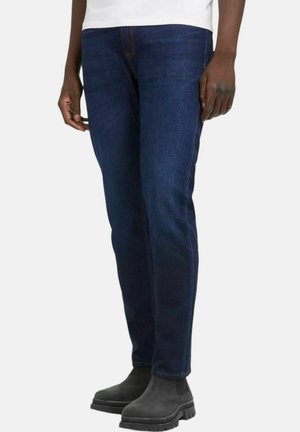 Jeans in denim blu scuro con design a gamba dritta, caratterizzati da dettagli di cucitura arancione e una texture liscia, abbinati a stivali alla caviglia grigi.