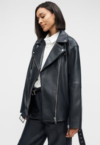 Γυναίκα με ίσια σκούρα μαλλιά που φοράει μαύρο δερμάτινο μπουφάν μοτοσικλετιστή, λευκό πουκάμισο με γιακά, κολιέ από μαργαριτάρια και μαύρο παντελόνι με ζώνη.