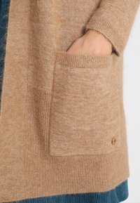 Cardigan de lana beige con una textura suave, que presenta un gran bolsillo frontal y puños de manga acanalados, mostrado sobre un fondo azul.