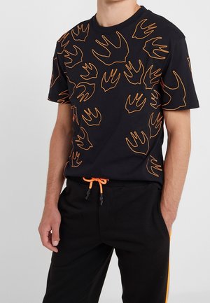 Man draagt een zwart T-shirt met korte mouwen met oranje handafdrukcontouren en zwarte broek met een oranje trekkoord en zijstreep.