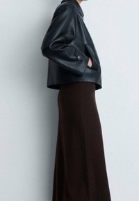 Schwarze Kunstleder-Cropped-Jacke mit übergroßen Ärmeln und einer einzelnen Fronttasche, kombiniert mit einem langen, eng anliegenden braunen Rock. Glatte Textur.