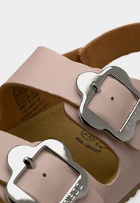 Κοντινό πλάνο των μπεζ δερμάτινων λουριών σανδαλιών με ασημένιες αγκράφες που φέρουν χαραγμένο το "FRIBOO", εμφανίζοντας τα ραφτά και μέρος του μαλακού πέλματος.