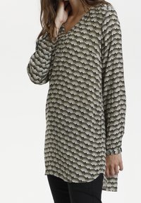 Tunique à manches longues en tissu vert foncé comportant un motif éventail en crème et noir, avec un décolleté en V et des fentes sur les côtés pour un ajustement décontracté.