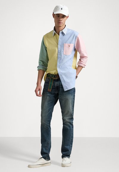 Camisa de botões multicolorida com seções em azul, verde, amarelo e rosa, com um bolso na frente. Combinada com jeans escuros e sapatos brancos.