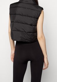 Schwarze ärmellose Daunenweste mit horizontalen Nähten, hohem Kragen und dezentem Branding auf der Schulter, kombiniert mit schwarzen gerippten Leggings.