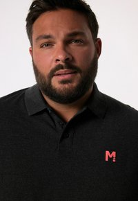 Černé polo tričko s krátkým límcem a dvěma knoflíky; na levé straně hrudi má červené logo. Tkanina se zdá být hladká a strukturovaná.