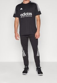Černý sportovní outfit Adidas obsahující tričko s krátkým rukávem, bílé logo a tři pruhy, spárováno s odpovídajícími kalhotami a černými teniskami.