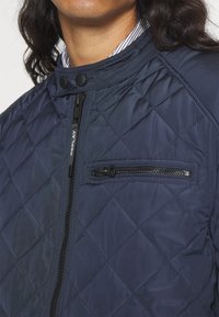 Homme portant une veste matelassée bleu marine avec une fermeture éclair verticale et une poche poitrine zippée horizontale sur un col de chemise rayée.