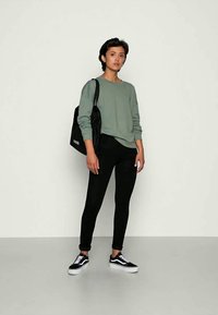 Sweatshirt verde claro, leggings pretas e tênis pretos usados por uma pessoa. Uma mochila preta com texto branco está pendurada em um dos ombros.