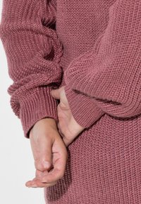 Sweter z dzianiny w kolorze rdzy, z prążkowanymi mankietami i fakturowanym ciałem. Ręce splecione z tyłu, podkreślając wzór dzianiny i miękki materiał.