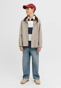 Jeune personne portant une veste beige avec un col foncé, un polo rayé bleu marine et blanc, un jean ample bleu, des chaussures beiges et une casquette rouge, regardant sur le côté.