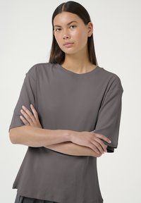 Grå kortærmet T-shirt med rund halsudskæring og afslappet pasform, der har en glat stof og et minimalistisk design. Modellen poser med korslagte arme.
