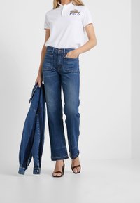 Vit polo-skjorta med broderad "POLO," kombinerad med högmidjade, raka blå jeans och bruna sandaler med remmar. Denimjacka hålls i handen.