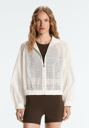 Donna che indossa una giacca bianca traforata con zip sopra una canotta aderente marrone scuro e pantaloncini, in piedi su uno sfondo chiaro uniforme.