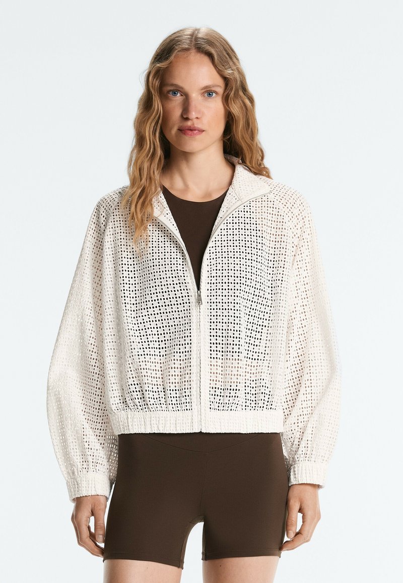 Femme portant une veste blanche perforée à fermeture éclair par-dessus un top moulant marron foncé et un short, debout devant un fond clair uni.