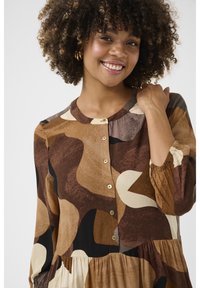 Barna és bézs mintás ruha hosszú ujjakkal, kerek dekoltázzsal és gombos előlappal. Az anyag lágy hatású, és folyékony vonalvezetésű.